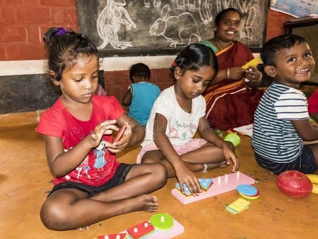 Young children sitting on the floor of a classroom playing with stacking and shape-sorting toys, with a smiling teacher behind them.
